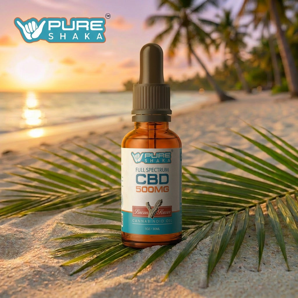 A bottle of Pure Shaka Full Spectrum CBD 500mg cannabis oil stands on a sandy beach with palm leaves in the foreground and a sunset ocean scene in the background.